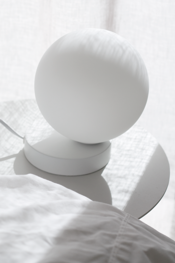 White globe table lamp on round bedside table with linen bedding in soft morning light.
