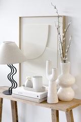Off-white speckled ceramic vase with sculptural curves styled on a console table with books, a black wavy lamp, and neutral accessories.