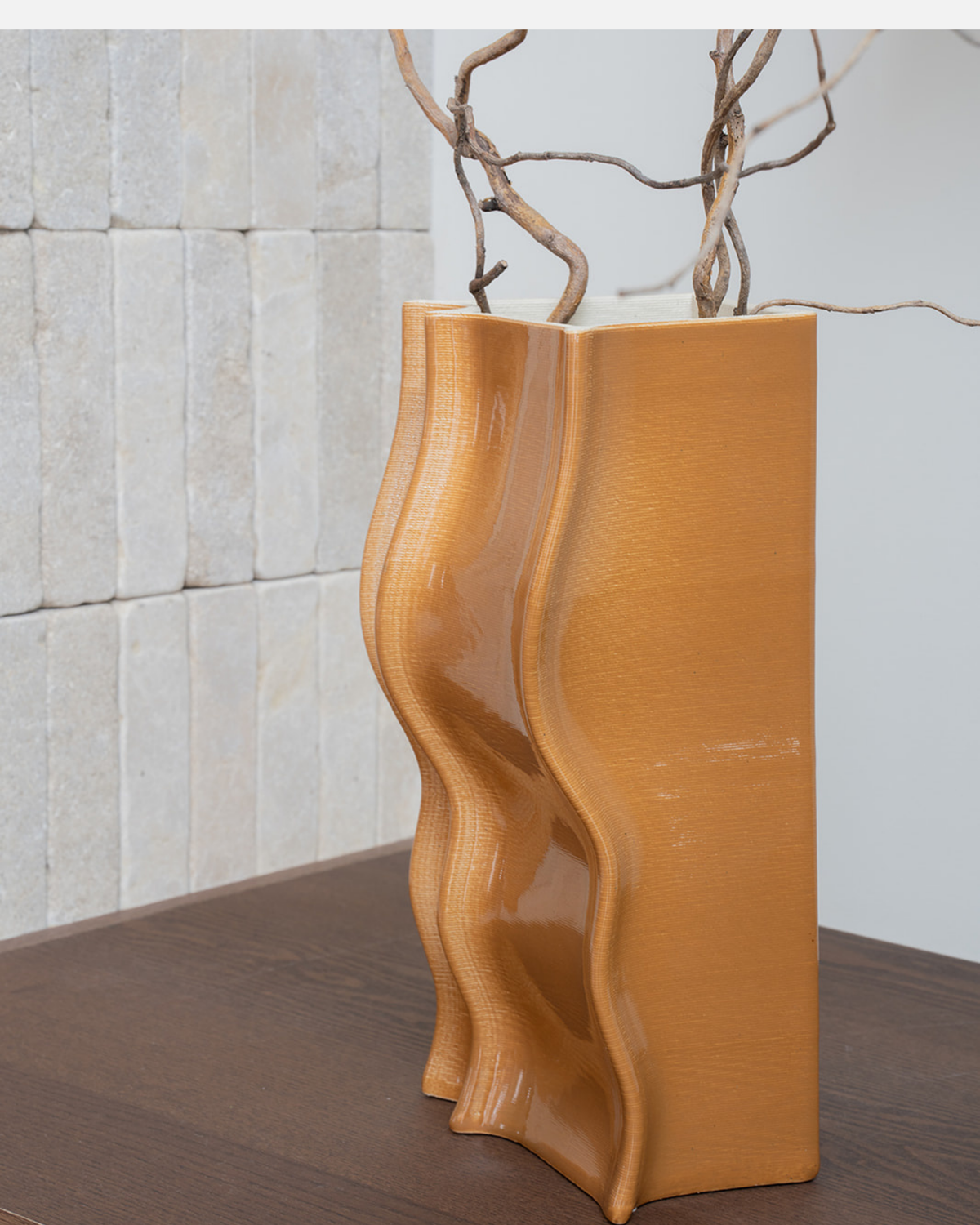 Close view of the vase in a glossy amber glaze holding twisted branches against a pale brick wall
