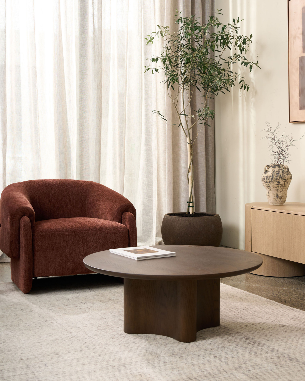 Umber armchair beside a potted olive tree and a round dark timber coffee table