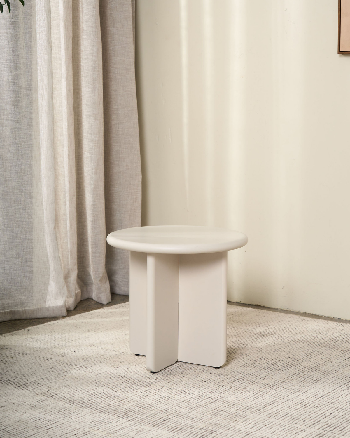 Cream side table with a cross base on a pale rug beside linen curtains