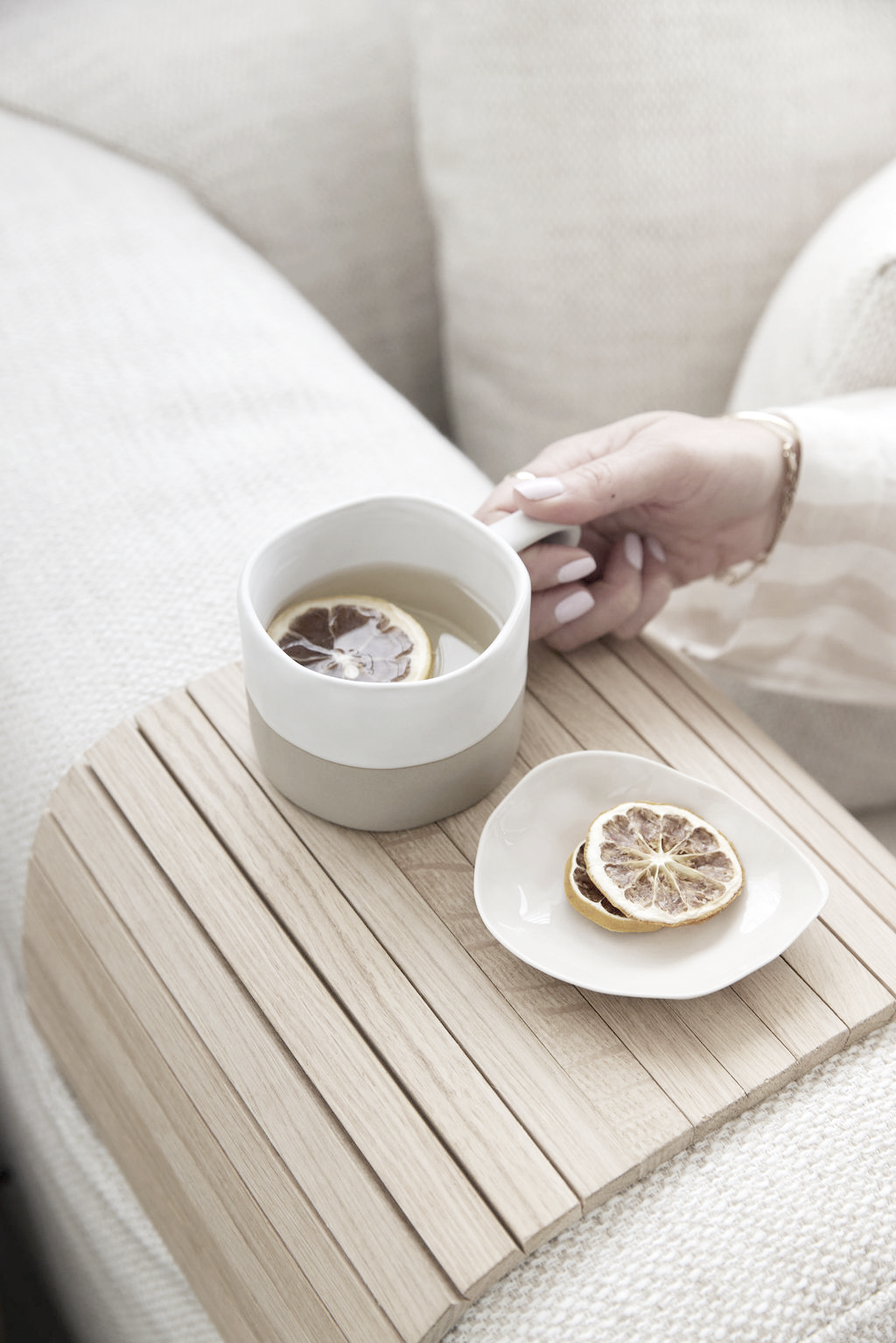 Hand reaching for a mug on the curved oak armchair tray, beside a small matching plate on a textured sofa