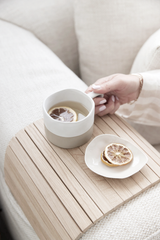 A woman’s hand reaches for a ceramic mug with dried lemon on a curved oak armchair tray, resting on a neutral textured sofa alongside a small matching plate.