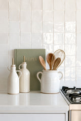 Olive linen Write To Me recipe journal styled on a white kitchen bench with ceramic oil bottles and a utensil crock against glossy white tiles.