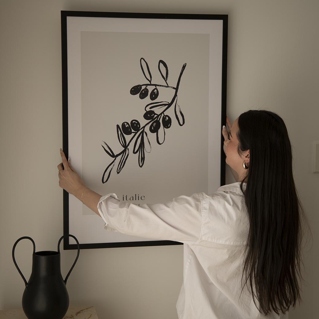 Woman hanging the Olive Tree print in a black frame above a black vase