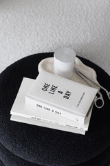 One Line a Day five year memory journal in neutral canvas cover stacked with books on a black boucle ottoman beside a stone tray with a white candle and wick trimmer
