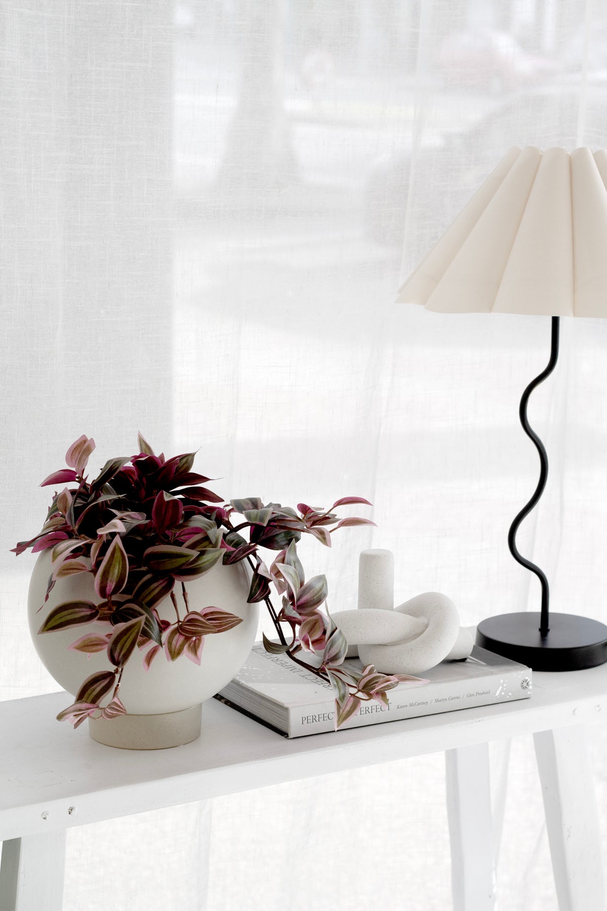 An oatmeal stoneware Orbit Planter with a trailing Tradescantia plant styled on a white console table alongside a wavy-stem table lamp, white ceramic objects, and an interior design book in a soft, light-filled Japandi-styled room.