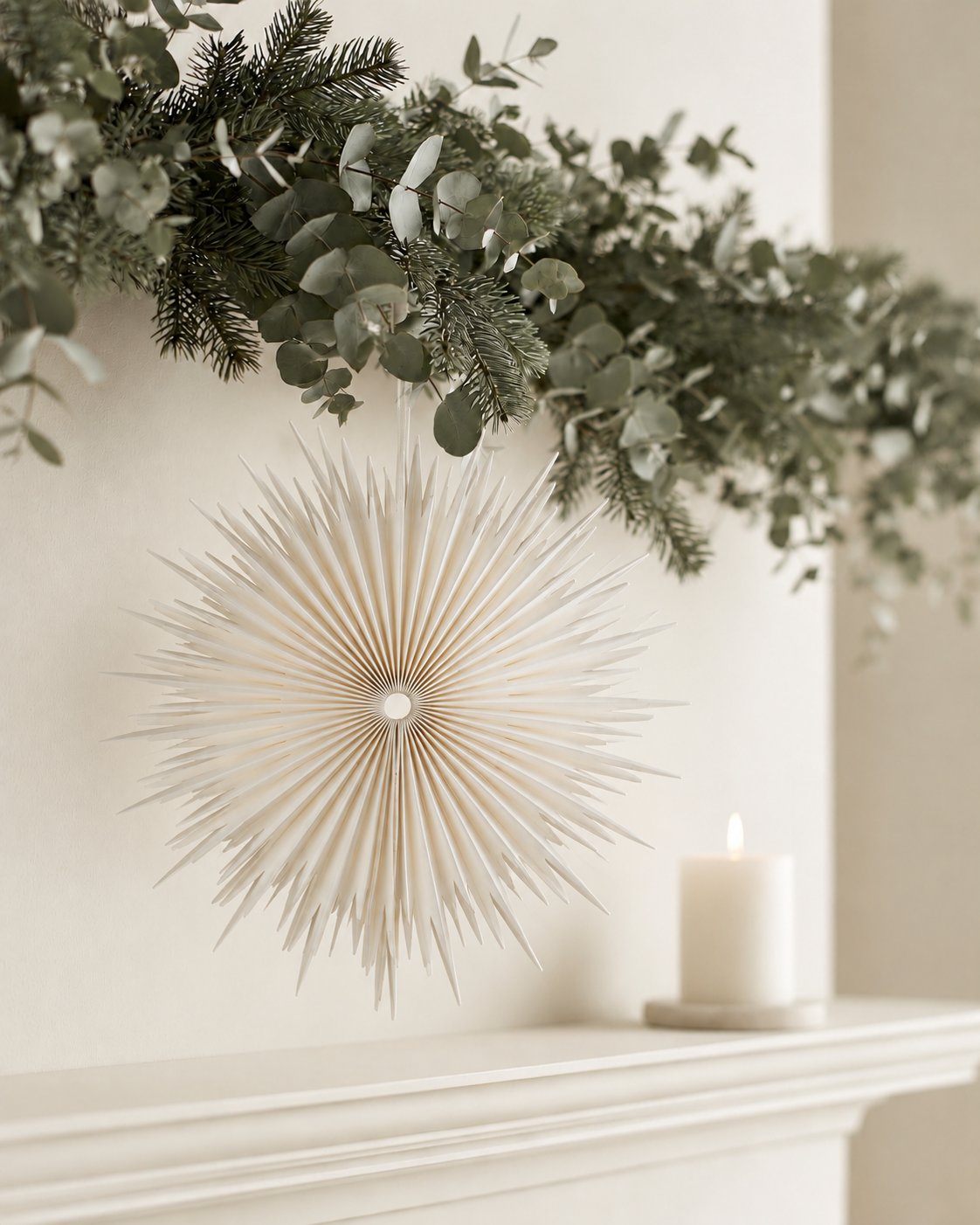 Off white paper snowflake hanging below a eucalyptus and pine garland above a mantel with a pillar candle