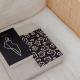 A lined black and white HOMEBODII notebook is sitting on top of a white pillow, perfect for jotting down notes and setting goals. (Brand Name: Papier HQ)