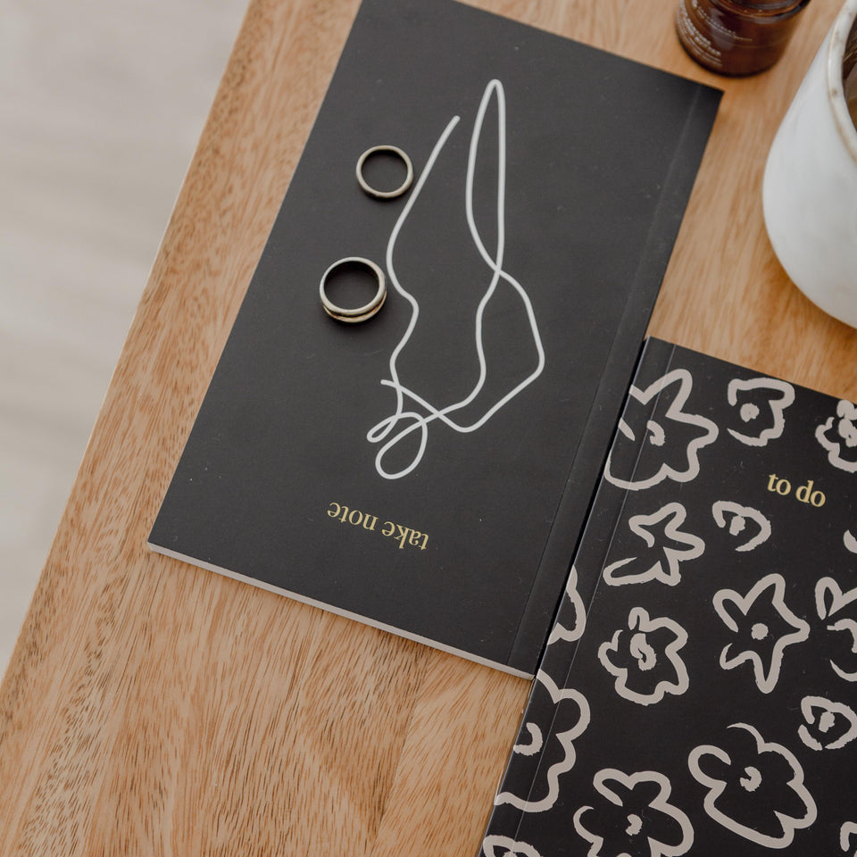 A HOMEBODII notebook from Papier HQ and a cup of coffee on a wooden table.