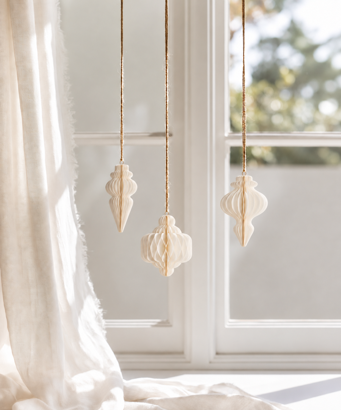 Three off white Papier gilded finials hanging on gold cords in front of a sunlit window with linen curtains