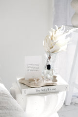 Papier HQ Travertine Curved Tray styled on a white side table with a wavy glass vase of dried white florals, gratitude print card, and The Art of Home book by Shea McGee, airy white bedroom setting.