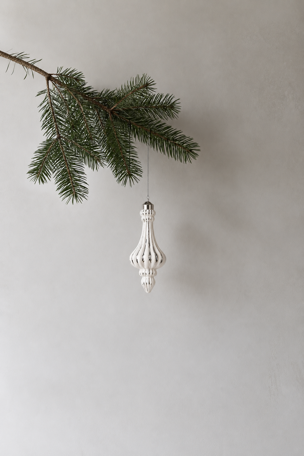 White and silver pendant bauble hanging from a pine branch against a soft grey wall