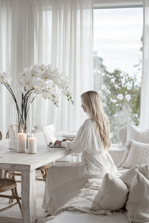 White faux Phalaenopsis orchid stem styled in a tall clear vase on a dining table with candles and soft neutral interiors.