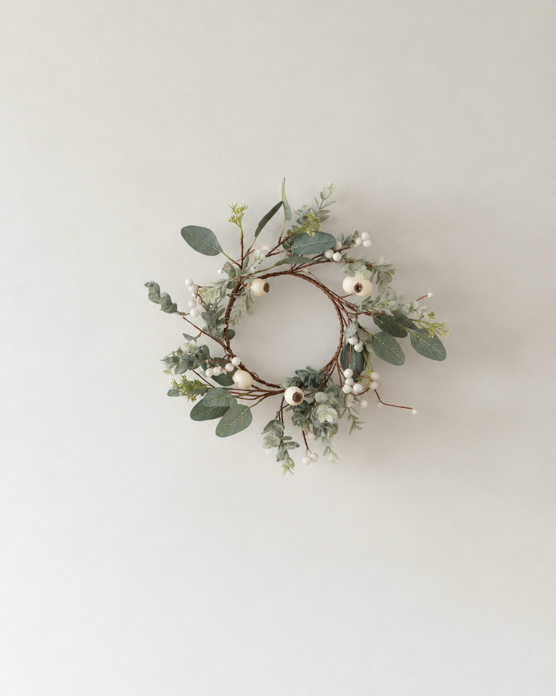 Podberry wreath in cream with sage leaves and cream pods hanging on a soft white wall