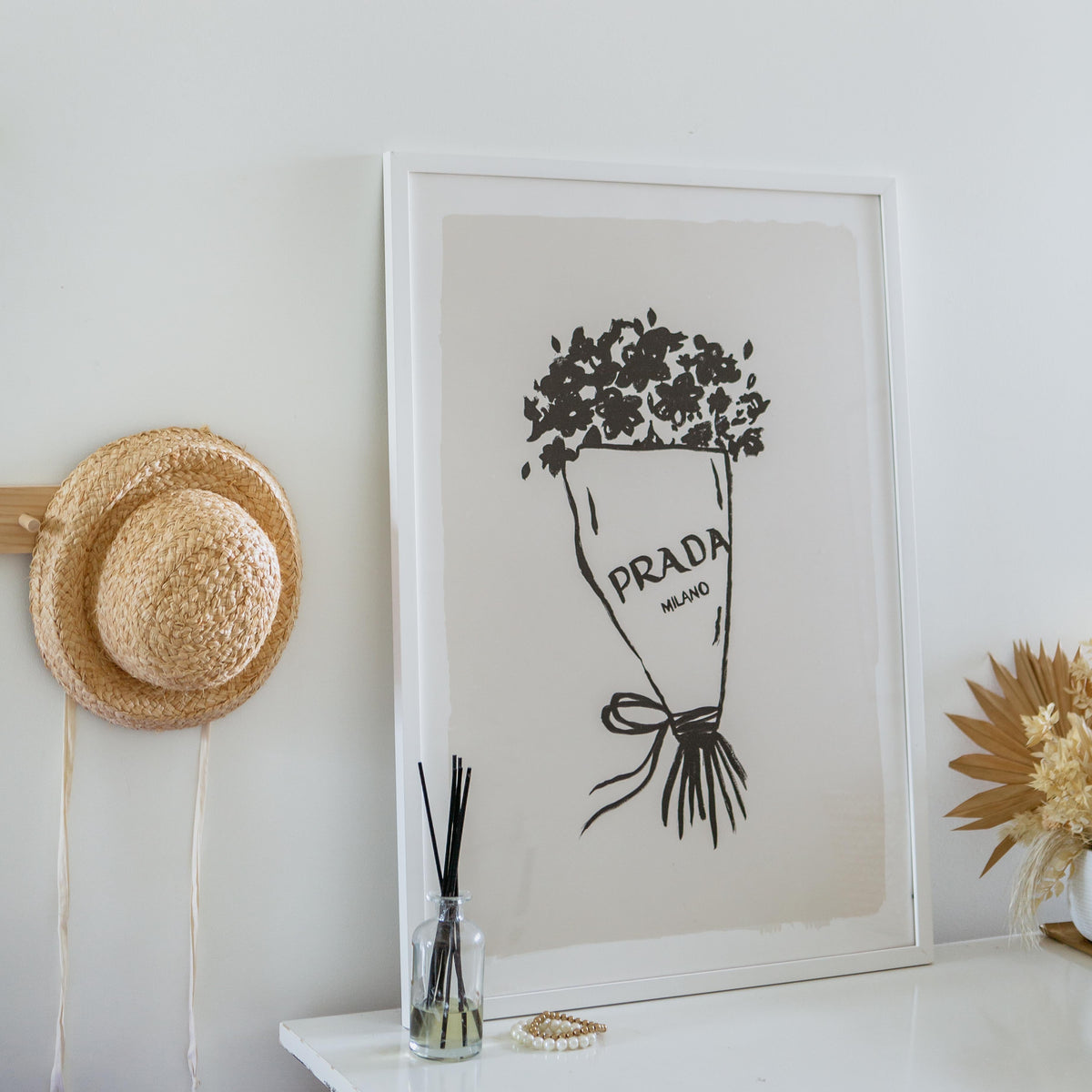 Prada Flowers print in a white frame on a white console with a straw hat and reed diffuser