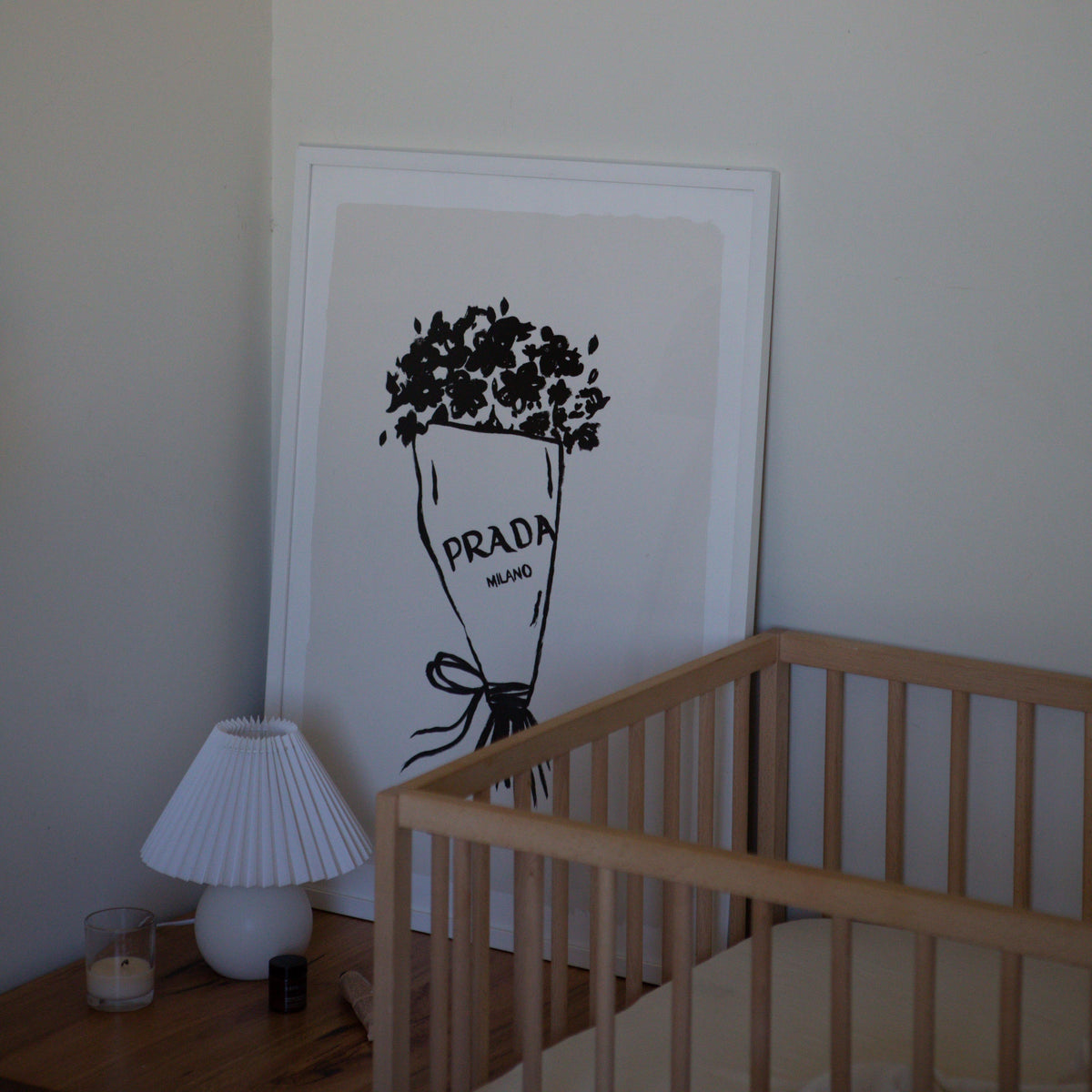 Prada Flowers print in a white frame leaning against a wall beside a timber cot and lamp