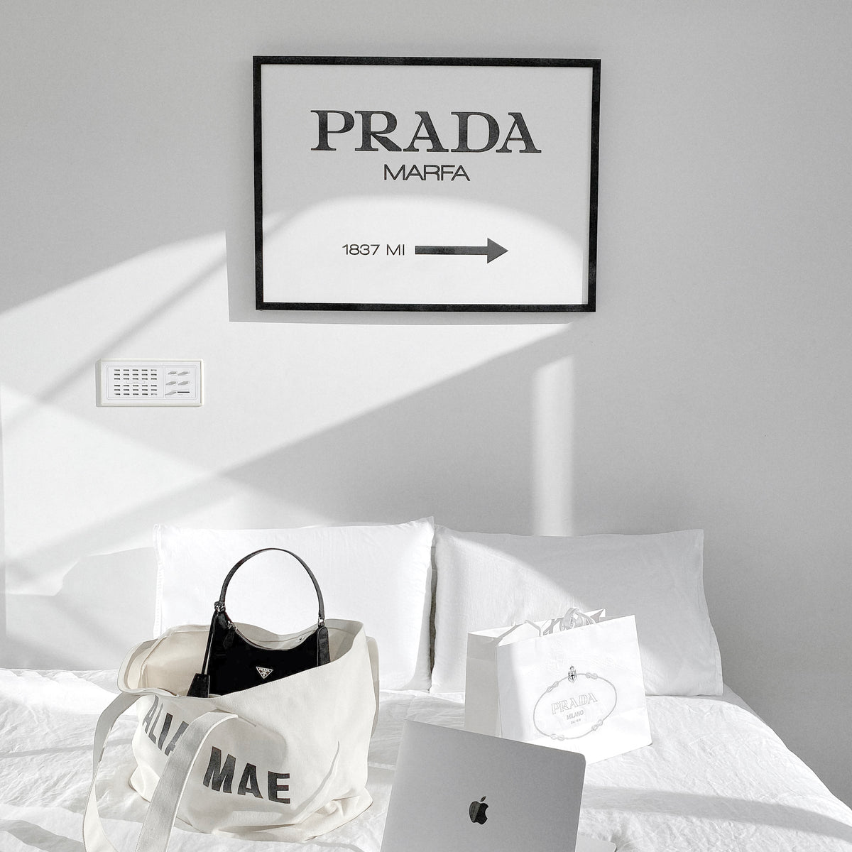 Prada Marfa print in a black frame above a bed with a tote bag and laptop