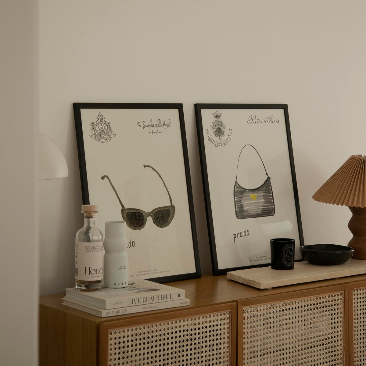 Prada Sunglasses and Prada Handbag prints in black frames on a rattan sideboard beside a pleated lamp