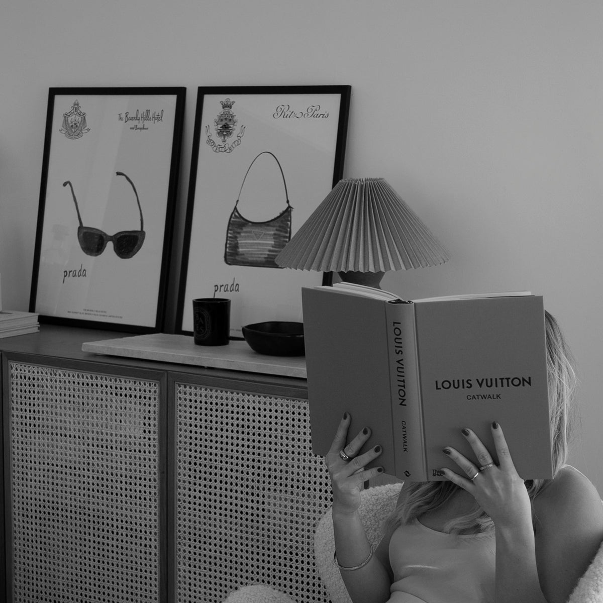 Black and white photo of Prada Sunglasses and Handbag prints on a rattan cabinet behind a woman reading