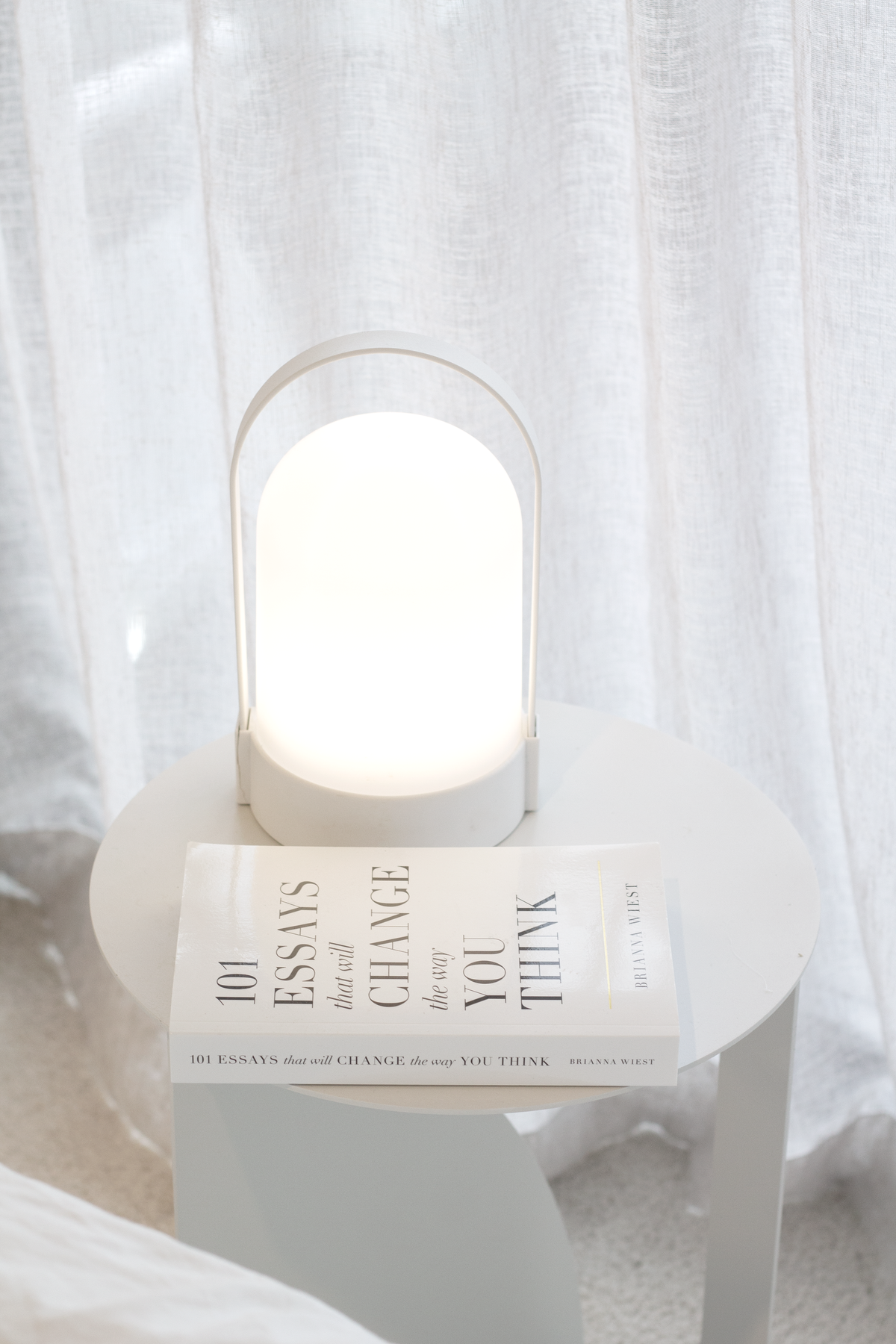 White portable LED lamp, unlit, on a stack of books in front of sheer curtains