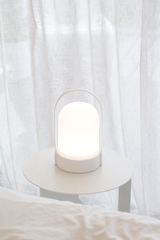 White rechargeable portable LED lamp glowing on round bedside table with sheer curtains