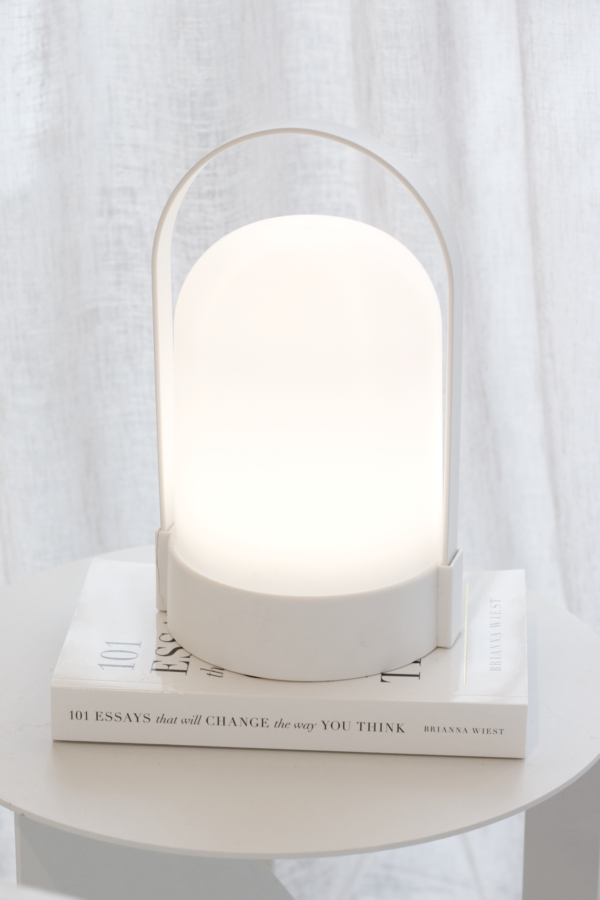 White portable LED lamp glowing on a book on a round side table in front of sheer curtains