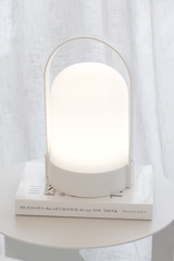Minimalist white portable LED lantern lamp with handle glowing softly on a round side table, styled on top of the book “101 Essays That Will Change the Way You Think” in front of sheer curtains.