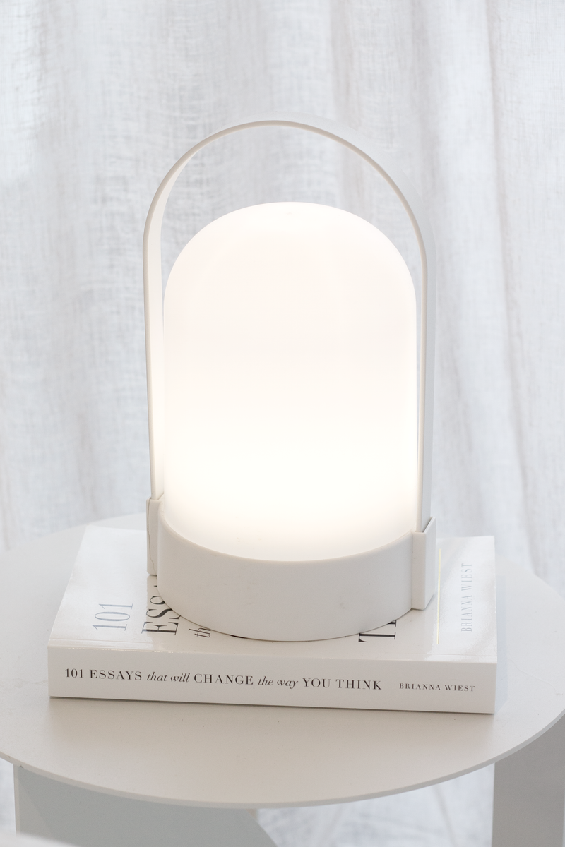 Minimalist white portable LED lantern lamp with handle glowing softly on a round side table, styled on top of the book “101 Essays That Will Change the Way You Think” in front of sheer curtains.