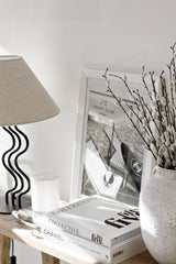 A set of soft faux pussy willow stems in a textured white vase, styled on a wooden console beside coffee table books, a sculptural black lamp, and a monochrome Chanel photo print.