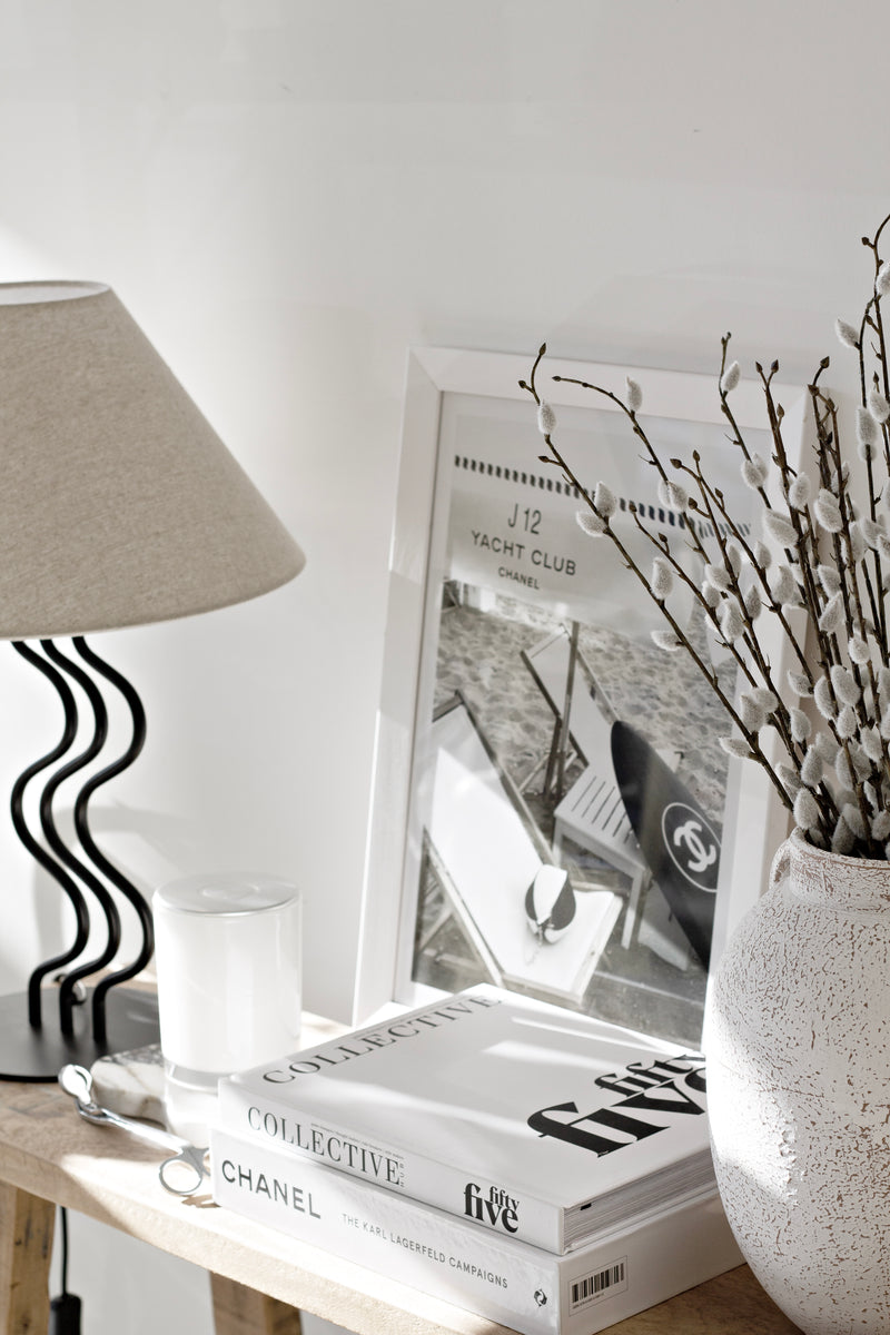 A set of soft faux pussy willow stems in a textured white vase, styled on a wooden console beside coffee table books, a sculptural black lamp, and a monochrome Chanel photo print.