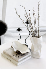 Bundle of tall faux pussy willow stems in a white sculptural vase on a round table, styled with stacked coffee table books, a wavy bowl and black candleholders.
