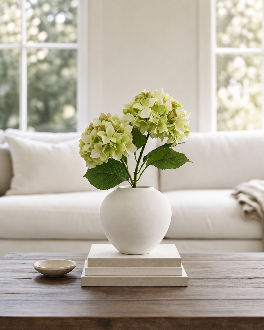 Two green Sublime Hydrangea stems in a round white vase on books on a timber coffee table