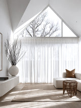 Natural rattan tulip stool beside boucle sofa with tan leather cushion in minimal white living room with vaulted ceiling and sheer linen curtains