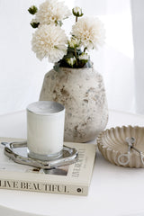 An elegant home decor vignette featuring realistic, aged-cream faux English Dahlia flowers arranged in a rustic, textured stone vase. The arrangement is styled on a white table with a "Live Beautiful" book, a silver candle tray with a white candle, and a concrete shell bowl, creating a serene and sophisticated scene.