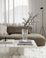Minimalist living room styled with a realistic faux magnolia bud branch in a ribbed glass vase on a marble coffee table layered with neutral books and a candle.

