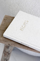 Oatmeal linen recipe journal with gold foil title on rustic wooden stool