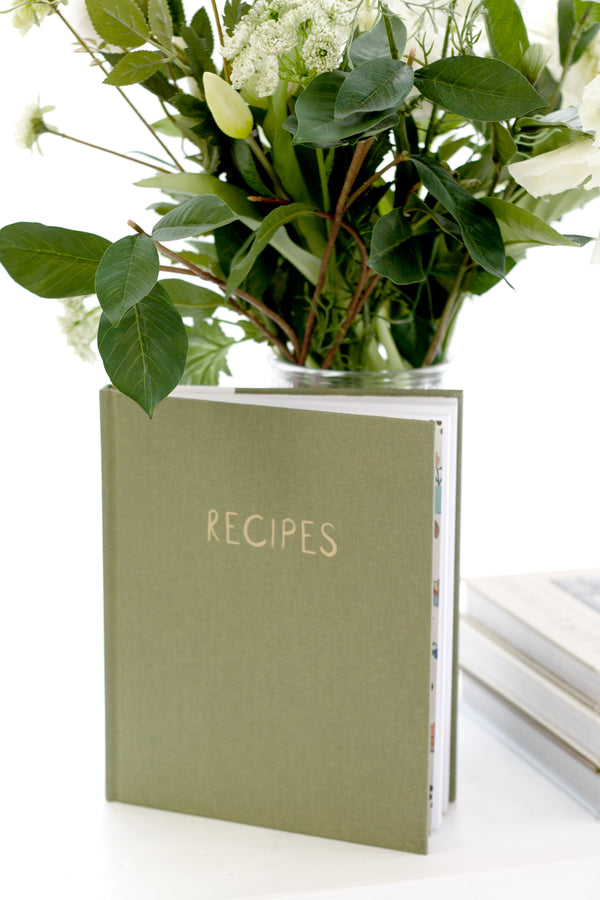 Olive green linen recipes journal standing upright with fresh white flowers behind, designed for recording favourite recipes and gifting to food lovers.