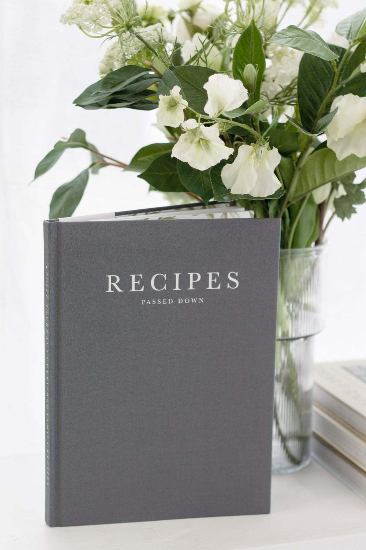 Recipes Passed Down journal in grey linen standing in front of a vase of white flowers