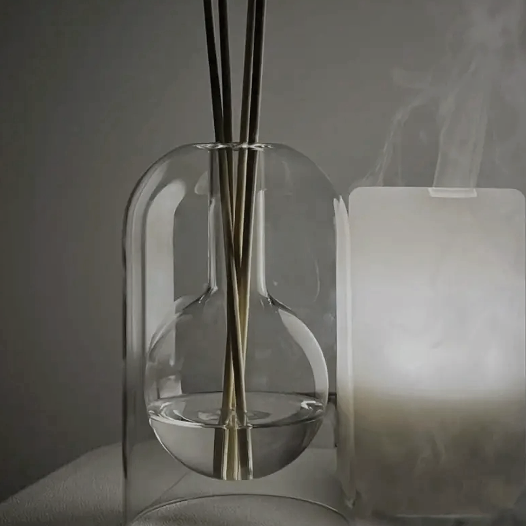 Glass reed diffuser with dark reeds beside a lit electric diffuser in a dim room