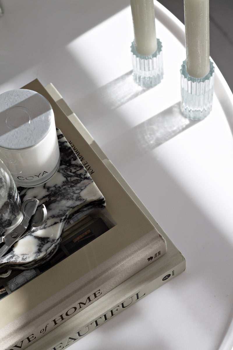 Pair of clear ribbed Ribb glass candle holders with white taper candles on a round white table, styled next to stacked coffee table books, a marble tray, an Ecoya candle and wick trimmer.