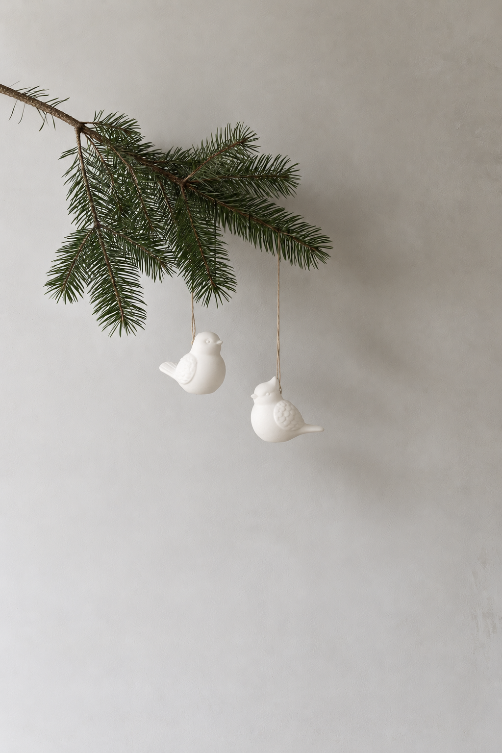 Two white robin hanging decorations on a pine branch against a soft grey wall