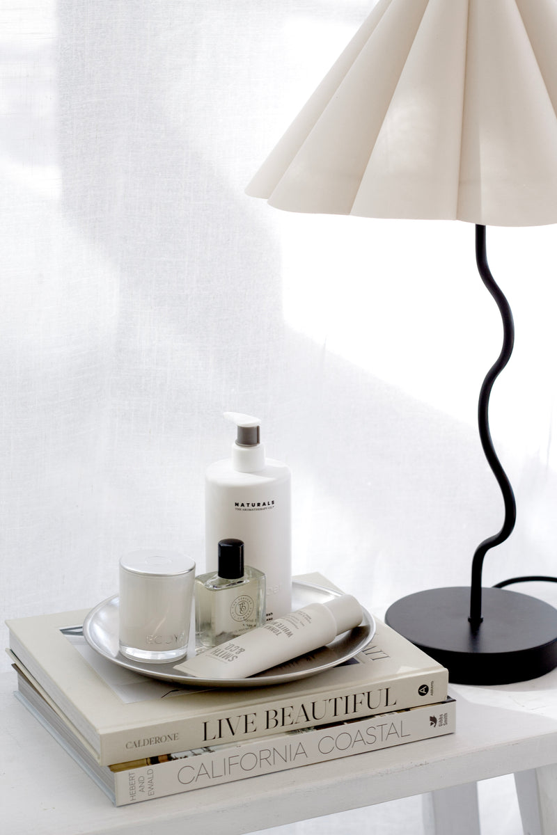 Silver round stainless steel decor tray styled on books with candle, perfume, skincare, and bedside lamp.
