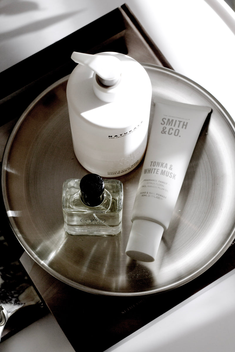 Round stainless steel tray in silver finish styled with lotion, hand cream and perfume; minimalist decor organiser.