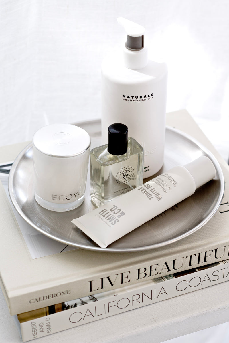 Silver round stainless steel tray styled on coffee table books with a candle, perfume, lotion and hand cream in soft light.