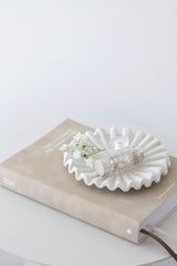 Glossy white fluted ruffle ceramic catchall tray holding small perfume vials and tiny white flowers, styled on a neutral diary on a round table