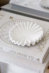 Glossy white ruffle ceramic display tray sitting on a linen-covered coffee table book titled Journeys, styled in soft neutral tones.