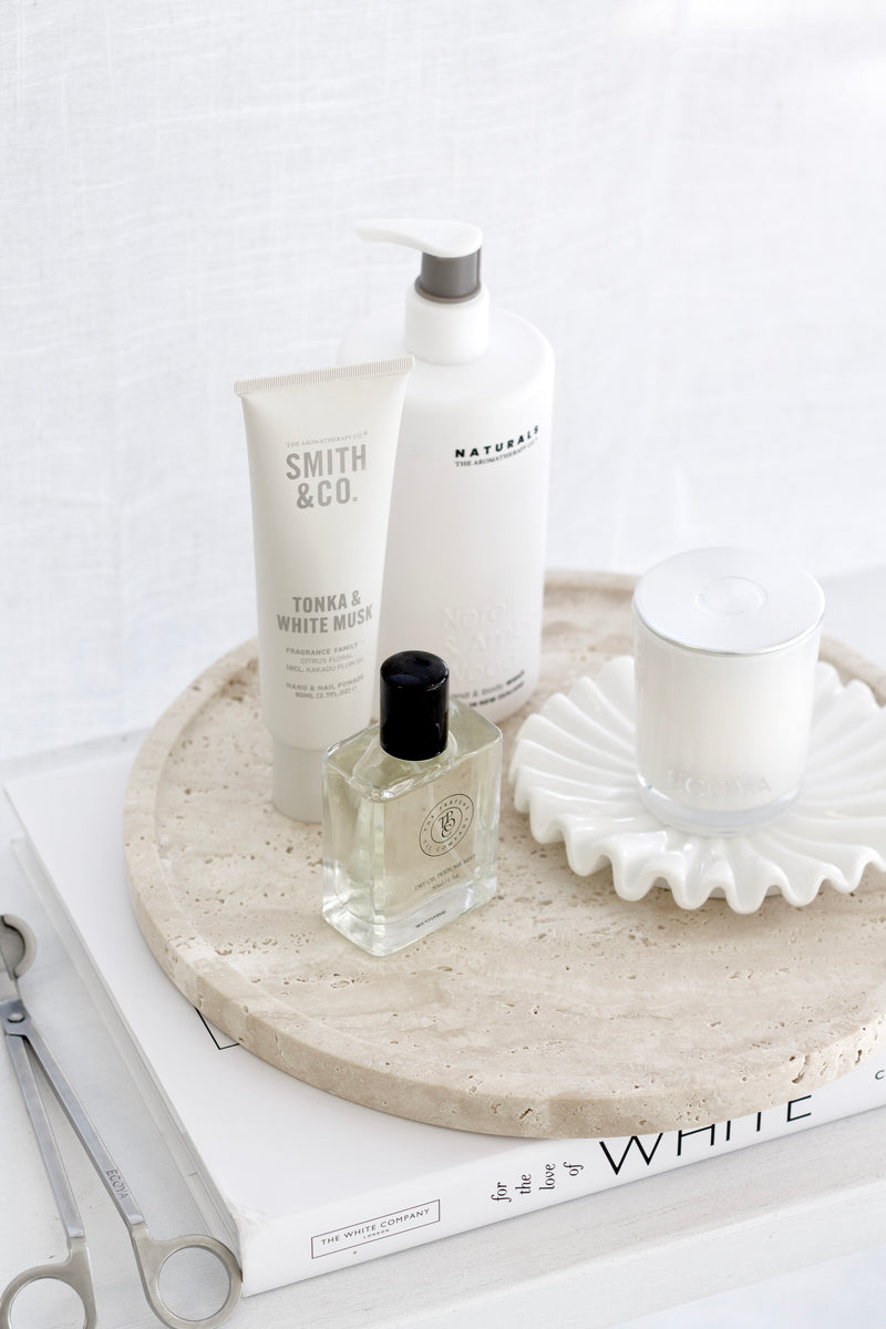 White ruffled ceramic tray styled with perfume, lotion, and candle on a travertine surface for a calm minimal look