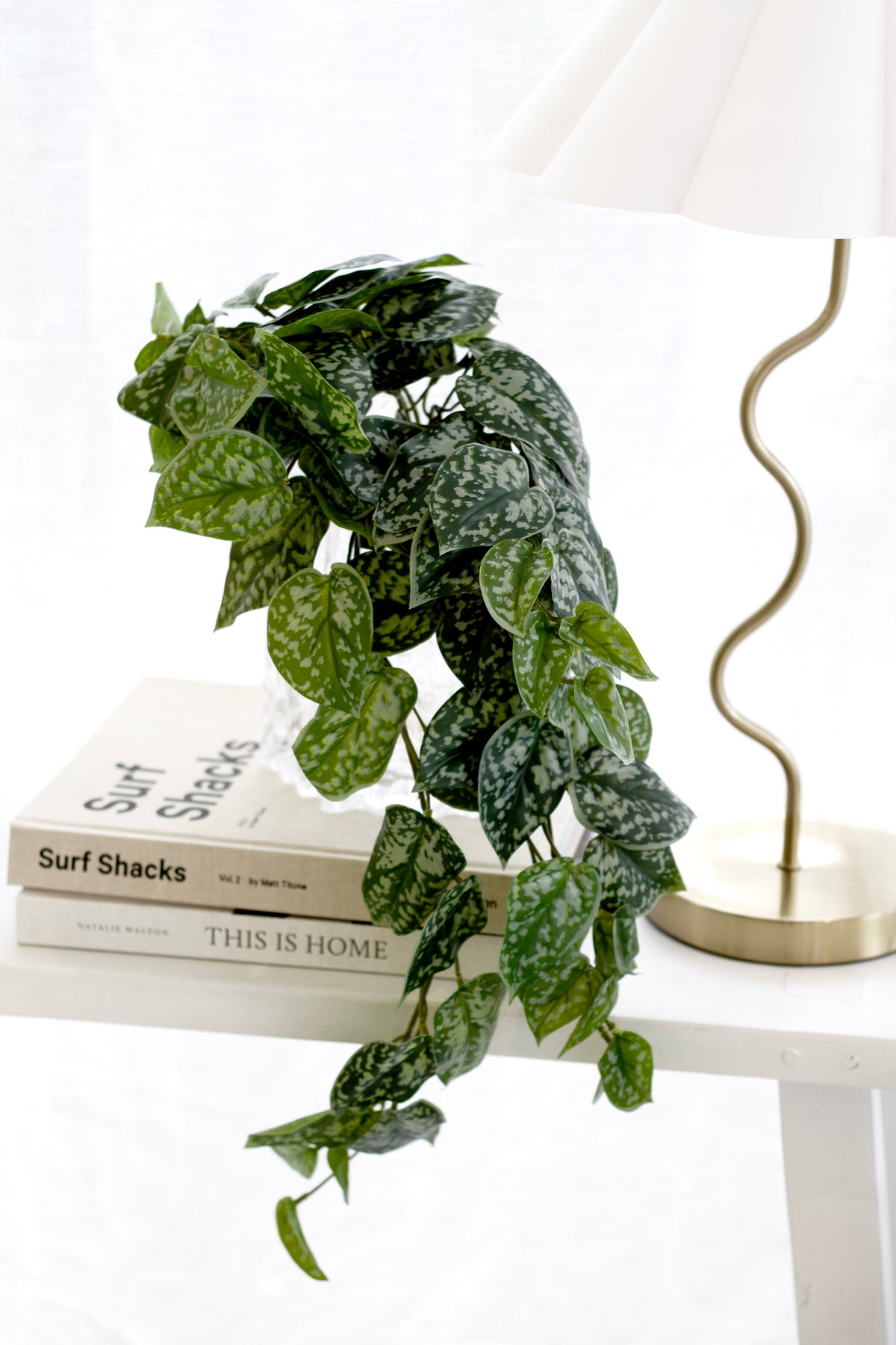Faux Scindapsus trailing from a white side table beside books and a wavy brass lamp