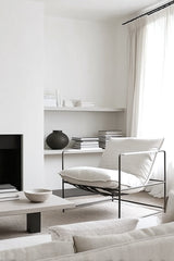 Minimalist white leather lounge chair with slim black iron frame in a bright neutral living room with shelves, linen curtains and a low stone table.